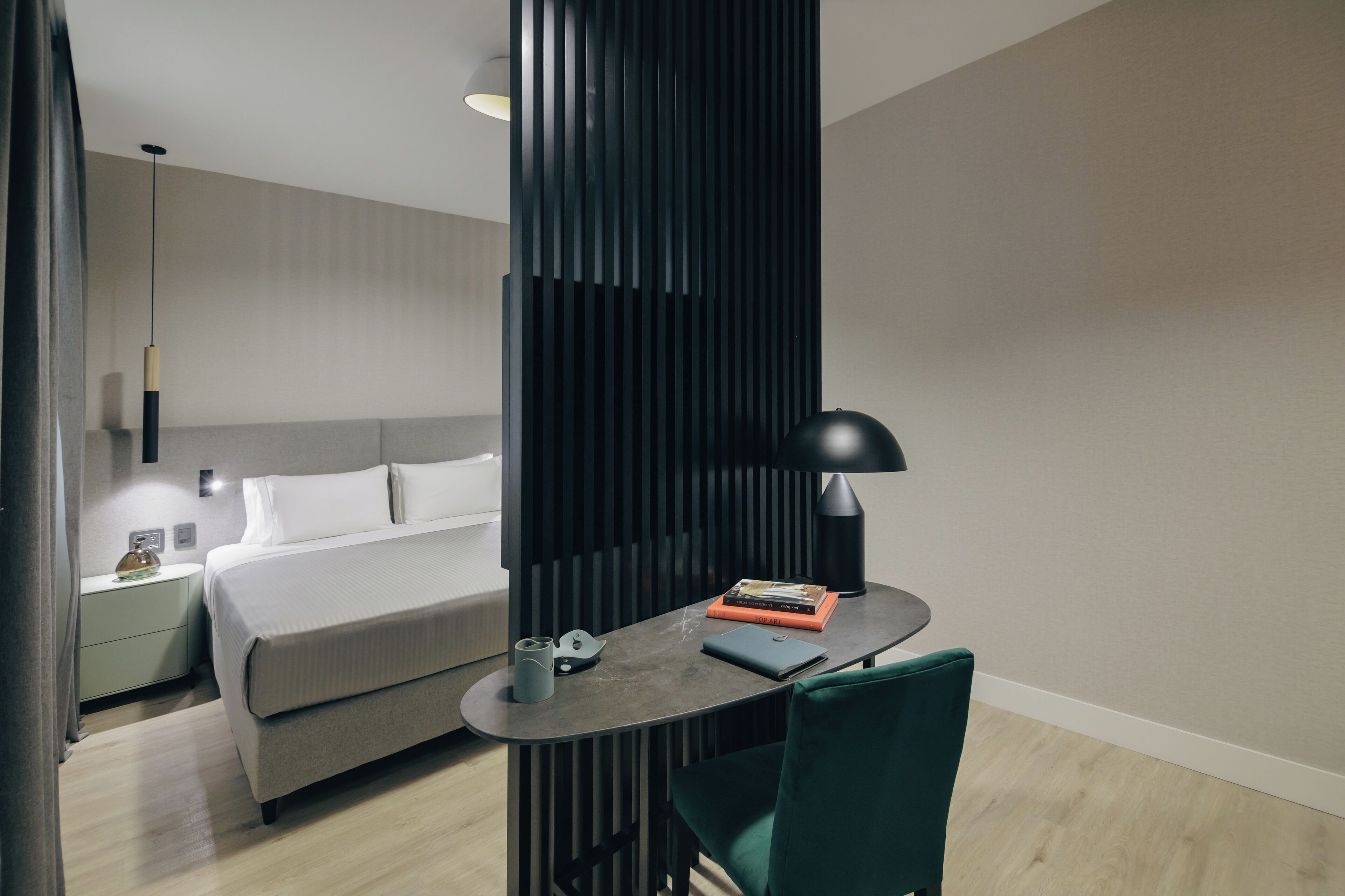 Una habitación de hotel moderna con cama, mesita de noche, lámpara, silla y un pequeño escritorio con lámpara y libros.
