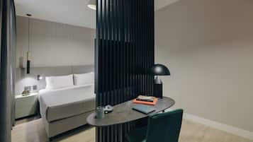 A modern hotel room with a bed, bedside table, lamp, chair, and a small desk with a lamp and books.