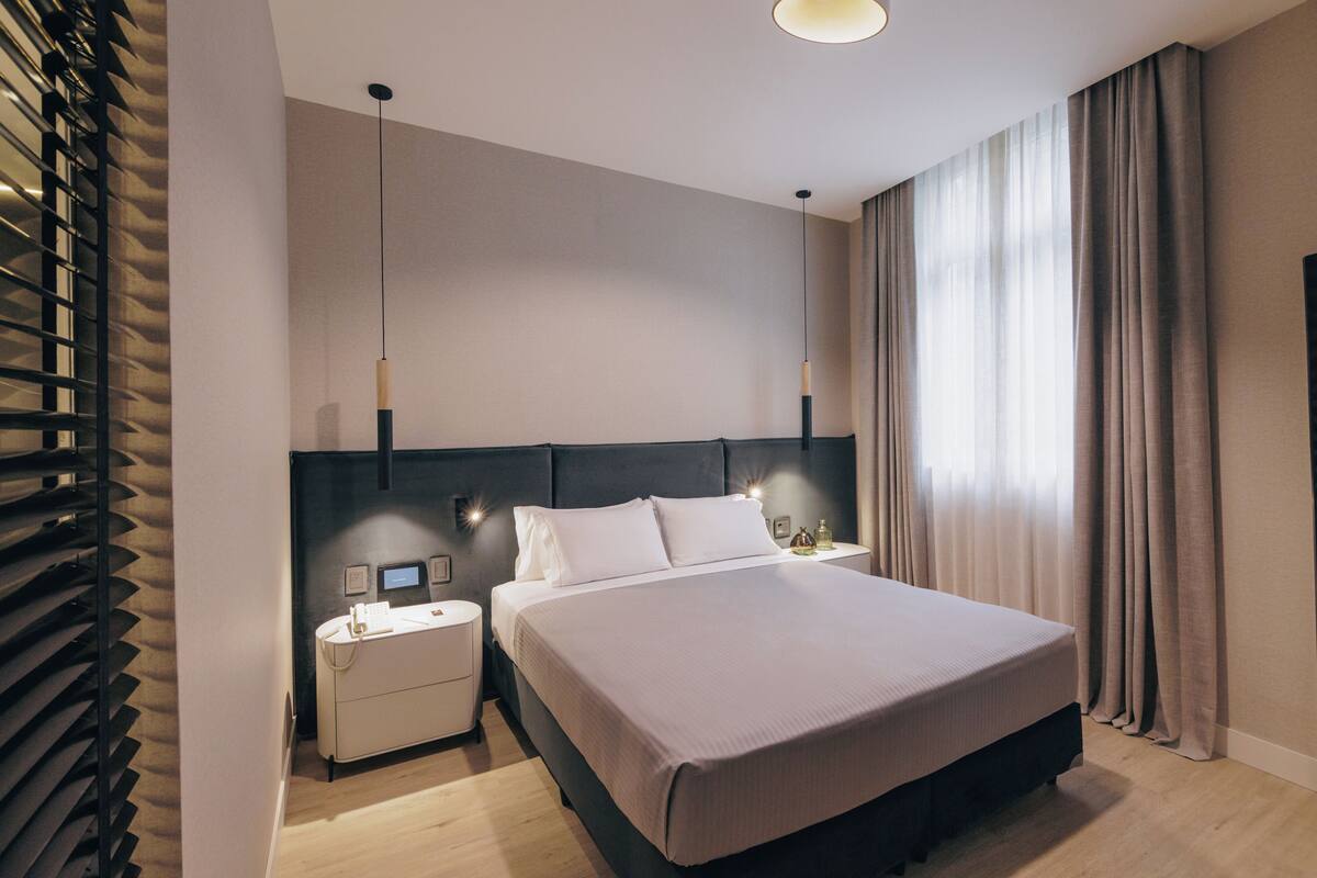 Una habitación de hotel moderna con una cama grande, mesitas de noche y un ventanal con cortinas.