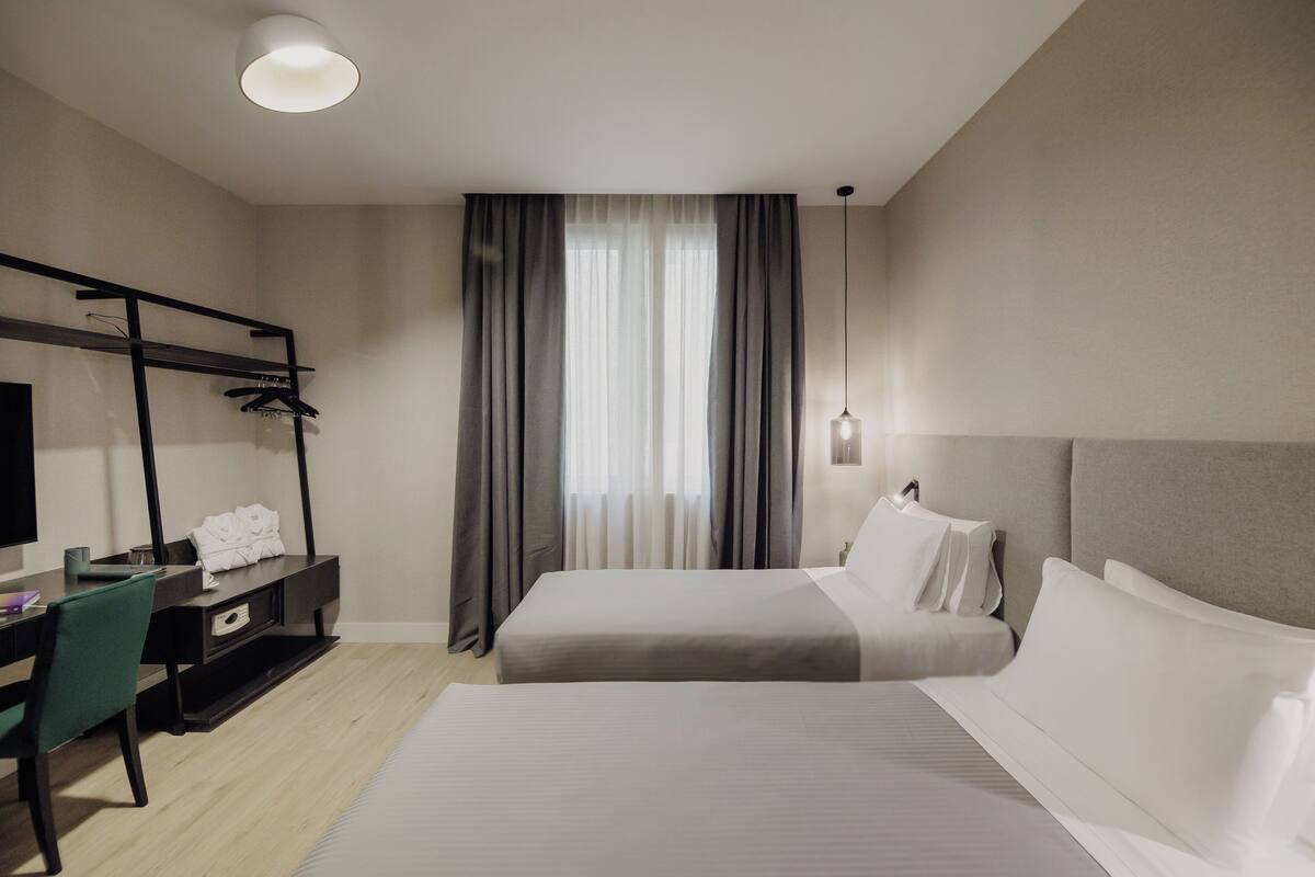 Una habitación de hotel con cama, escritorio, silla, armario y ventana con cortinas.
