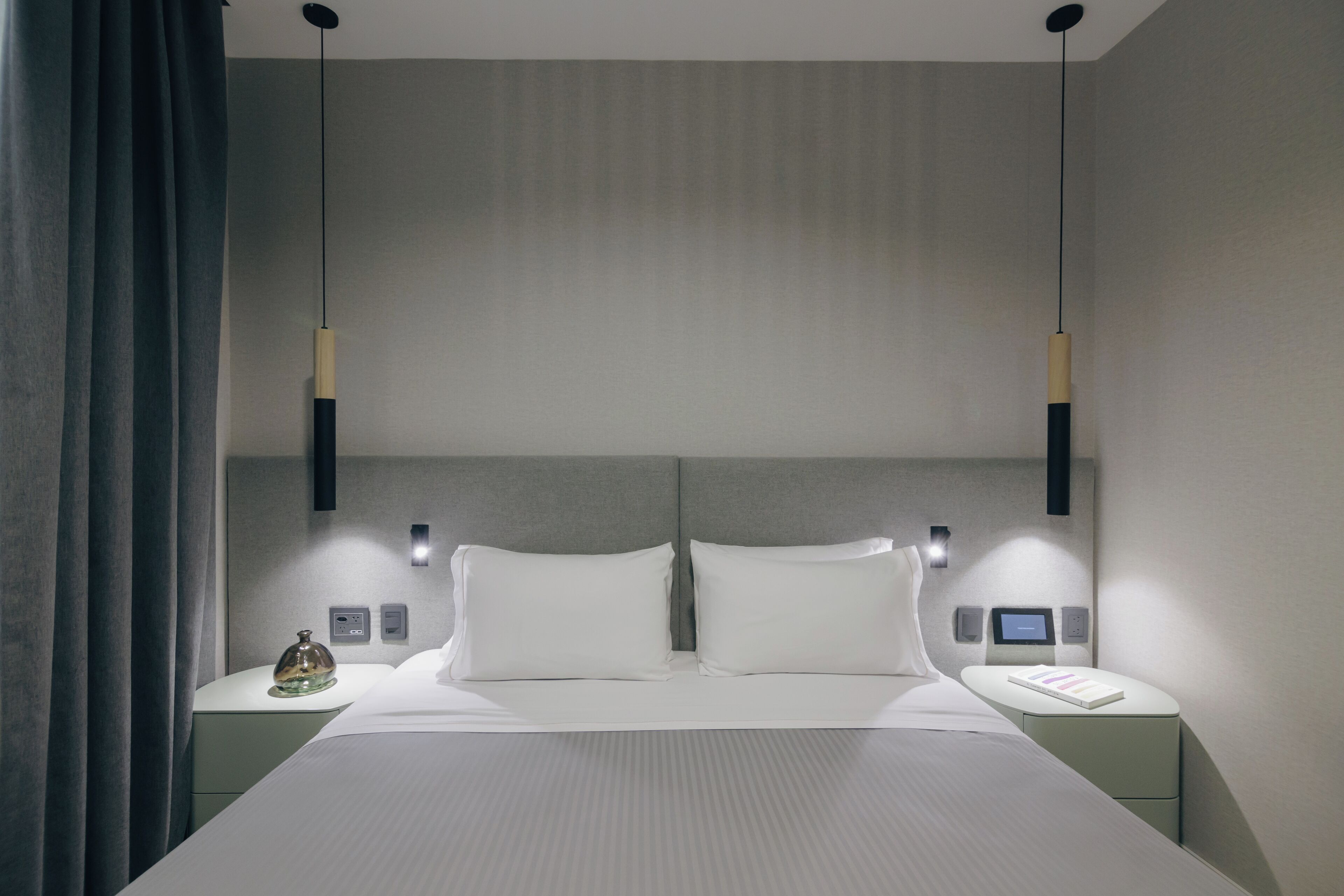A modern hotel room with a large bed, two bedside tables, a nightstand, a wall-mounted control panel, and a ceiling light fixture.