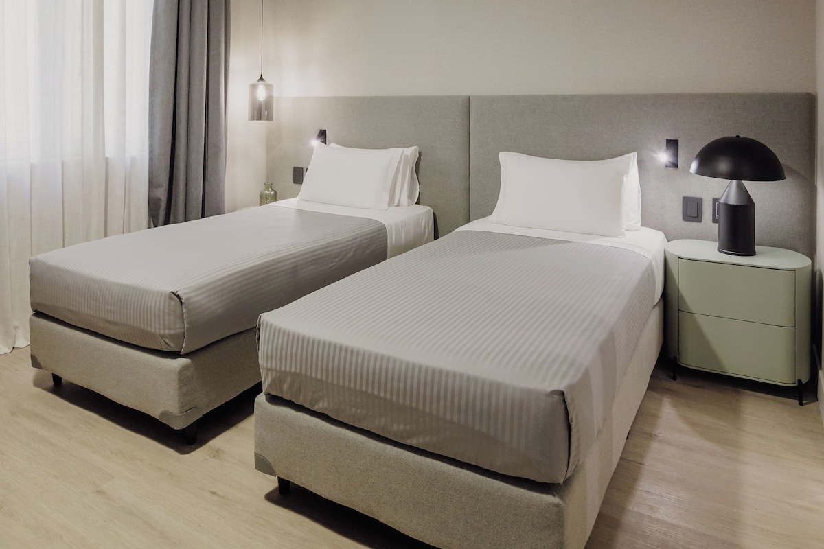 Una habitación de hotel moderna con dos camas, una mesita de noche con lámpara y un gran ventanal con cortinas.