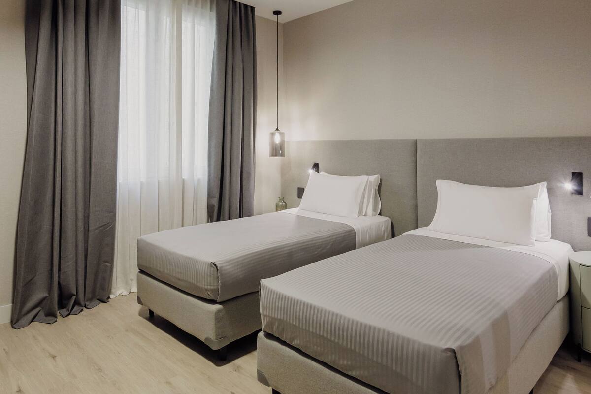 Habitación de hotel con dos camas, un ventanal grande con cortinas, una lámpara de techo y una mesita de noche con lámpara.