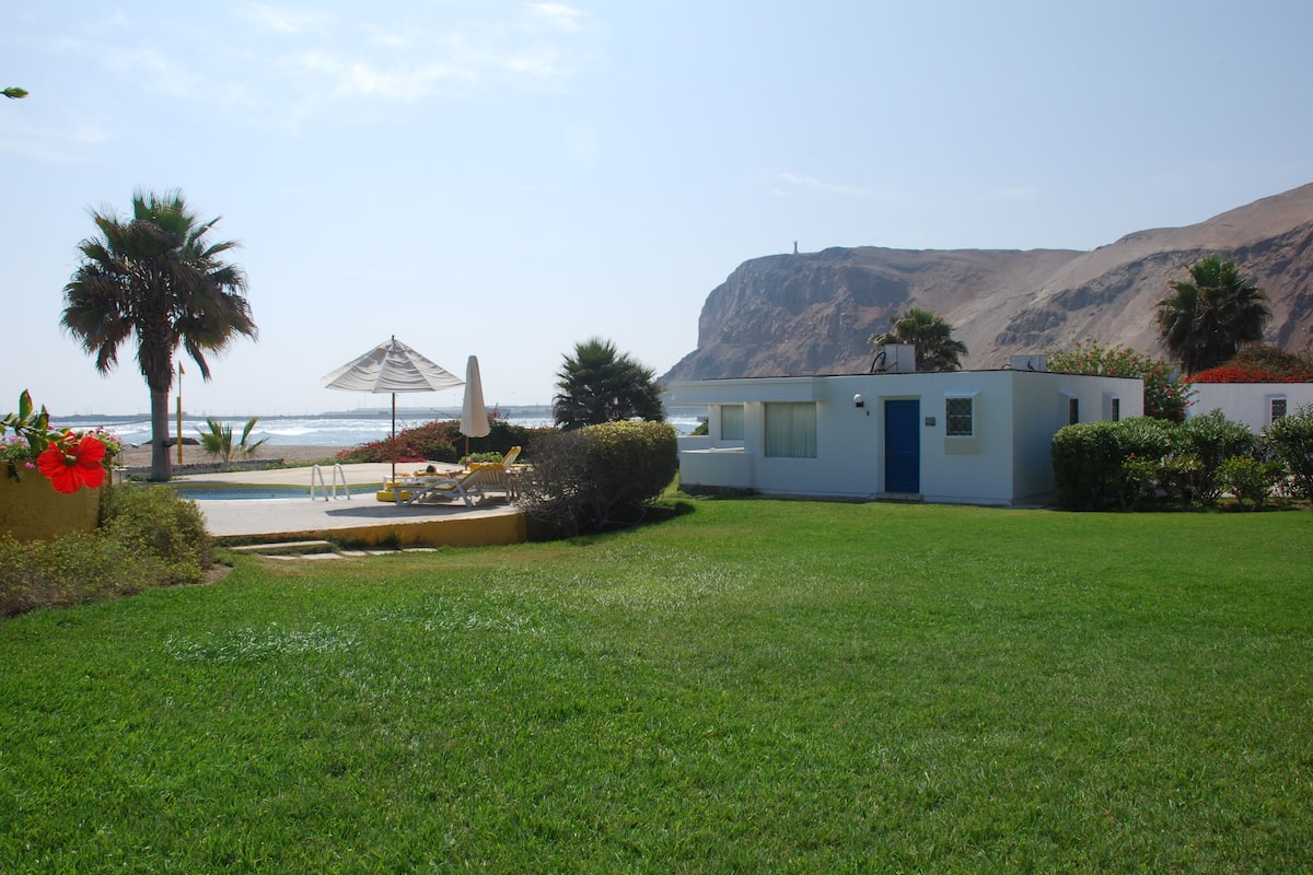 A small, single-story building with a blue door, situated in a grassy area with a pool and lounge chairs. A mountain is visible in the background.