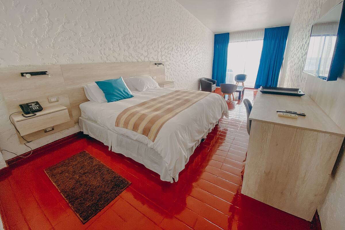A hotel room with a bed, bedside table, desk, chair, and a small window with curtains.