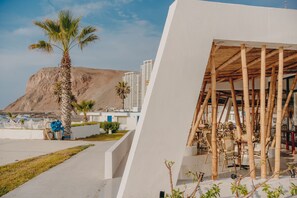 A modern building with a white facade, a bamboo structure with chairs, and palm trees in the foreground. - Hotel Arica (Arica)