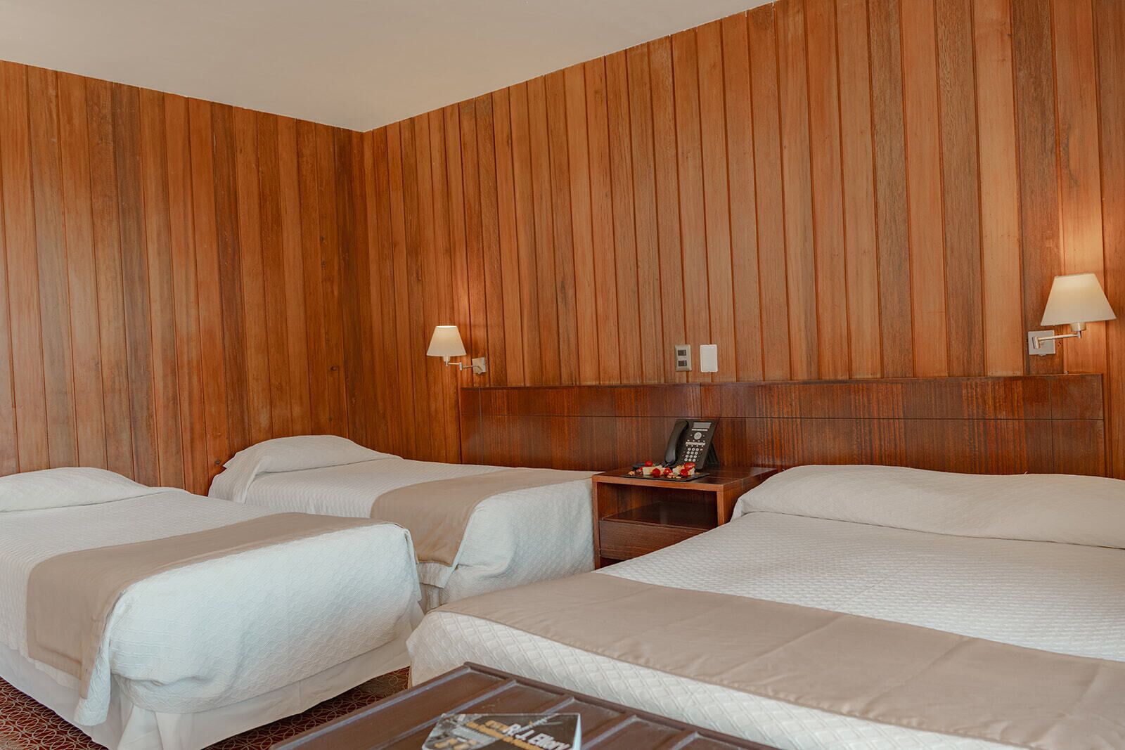 Habitación de hotel con tres camas, paneles de madera, lámparas de noche, teléfono y una mesita con una bandeja de frutas.