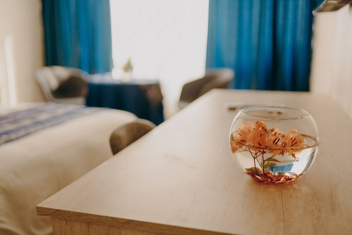 A hotel room with a wooden table, a glass vase with flowers, and a bed with a blue blanket.