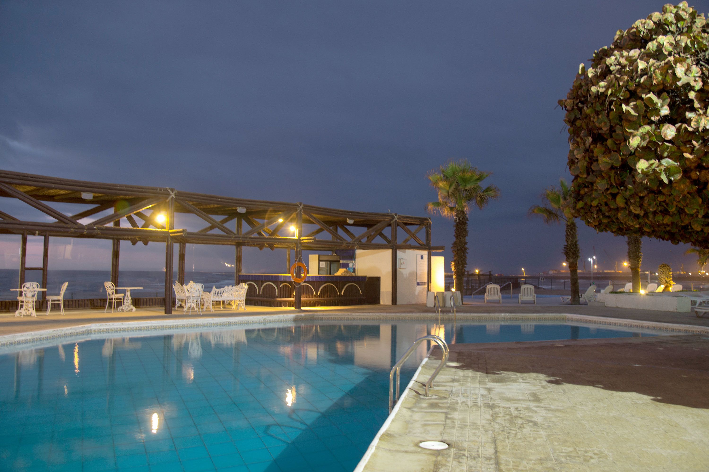 A pool area with a covered seating area, palm trees, and a view of the sea.