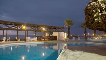 A pool area with a covered seating area, palm trees, and a view of the sea.