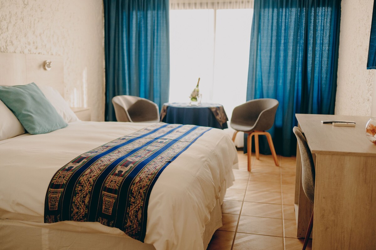 Habitación de hotel con cama, escritorio, dos sillas, una mesita con un florero y una ventana con cortinas azules.