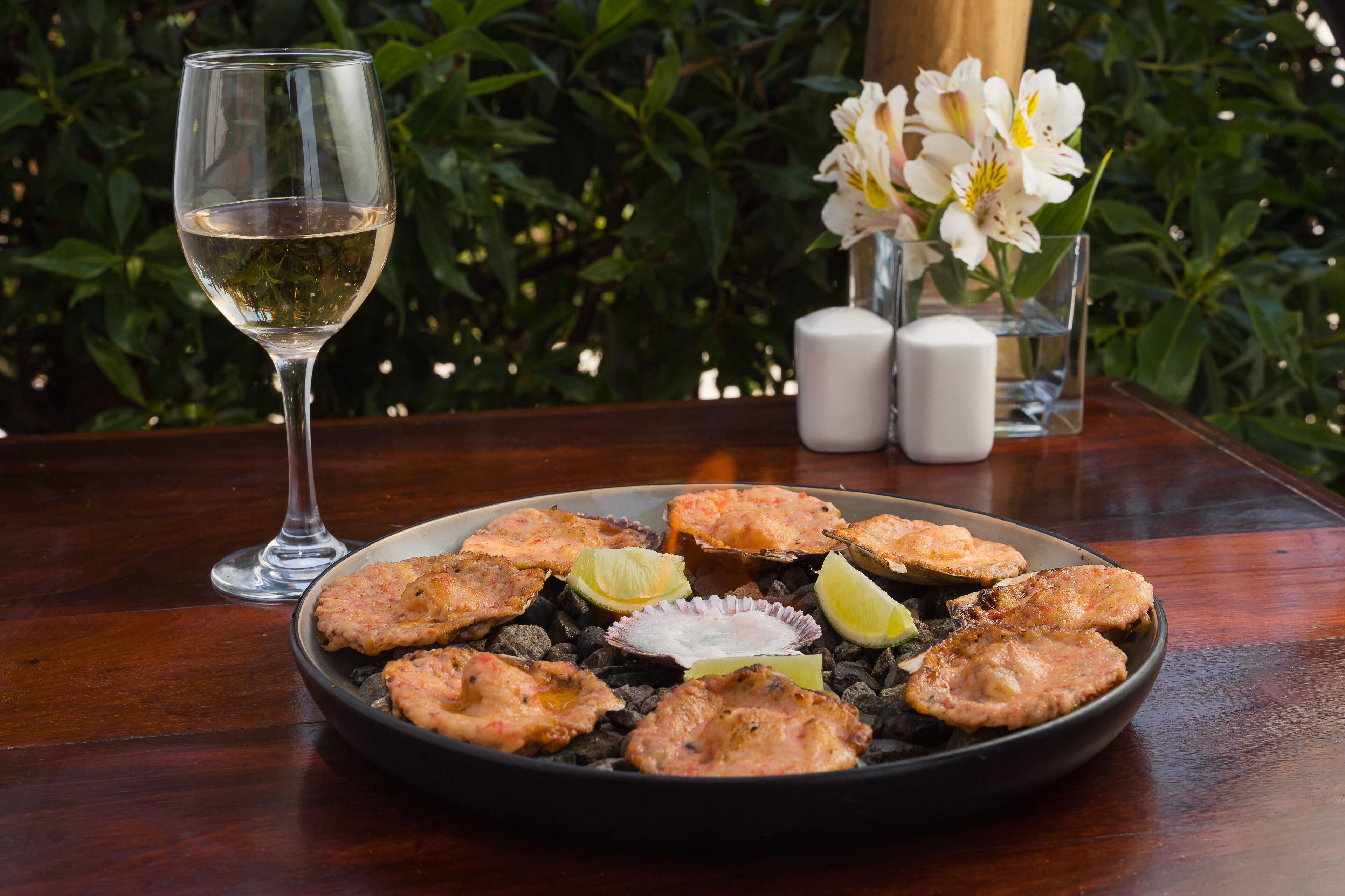 A plate of grilled fish with lime wedges, a glass of white wine, and a vase of white flowers on a wooden table.