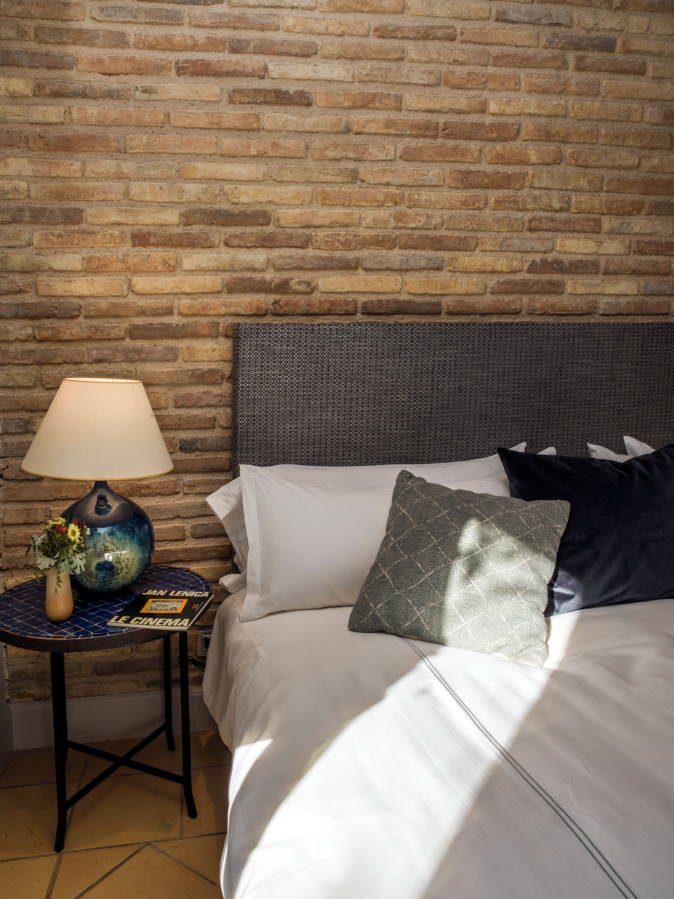 Une chambre d’hôtel avec un mur en briques, un lit recouvert de draps blancs, une tête de lit sombre et une petite table sur laquelle se trouvent une lampe et un vase de fleurs.
