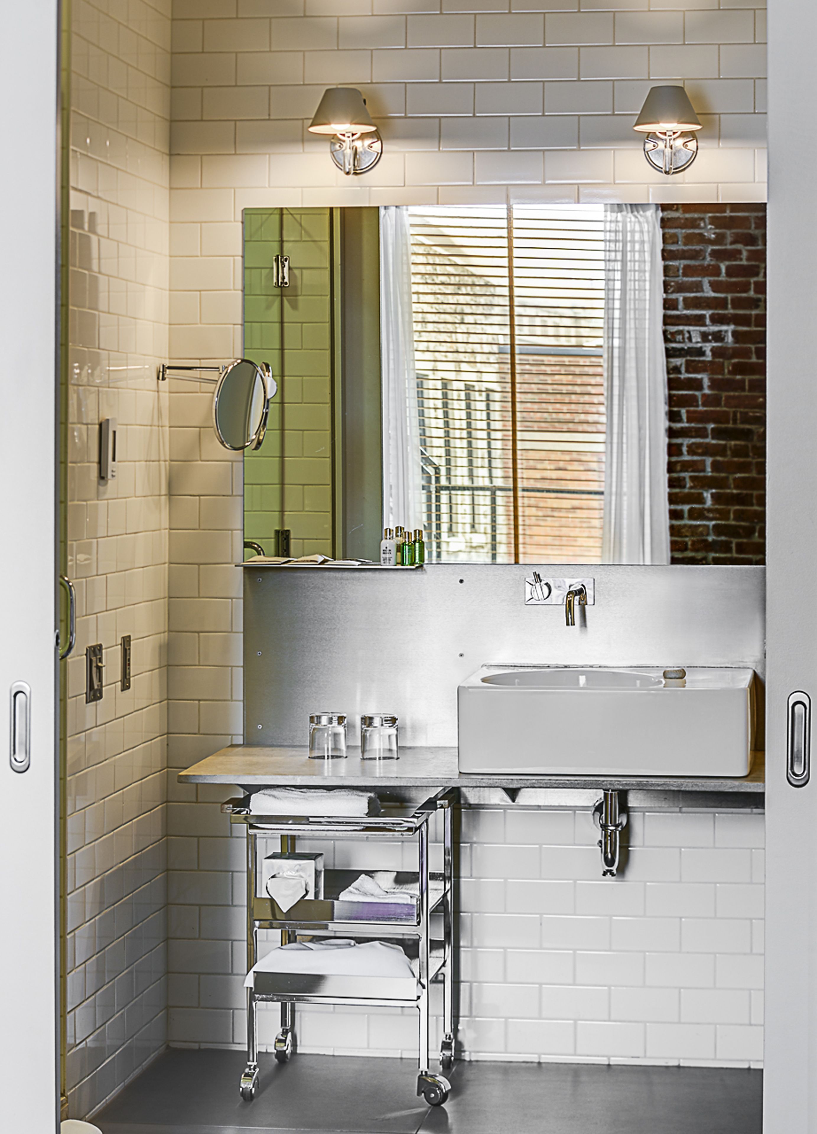 A modern bathroom with a white tiled wall, a large mirror, a sink, and a window with blinds.