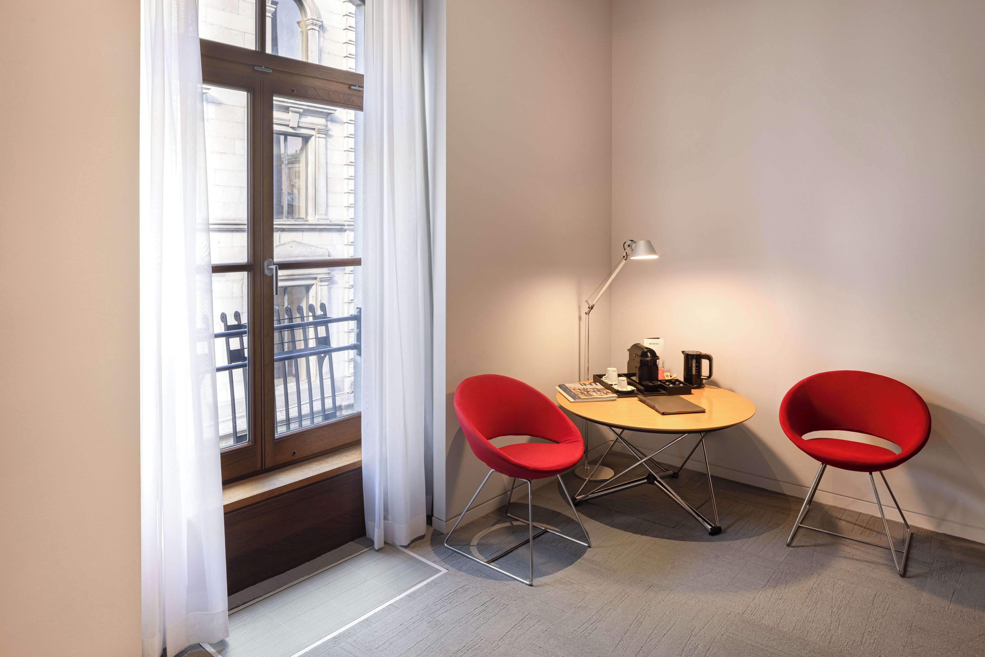 Une petite pièce avec une fenêtre, deux chaises rouges, une table avec une lampe et une machine à café.