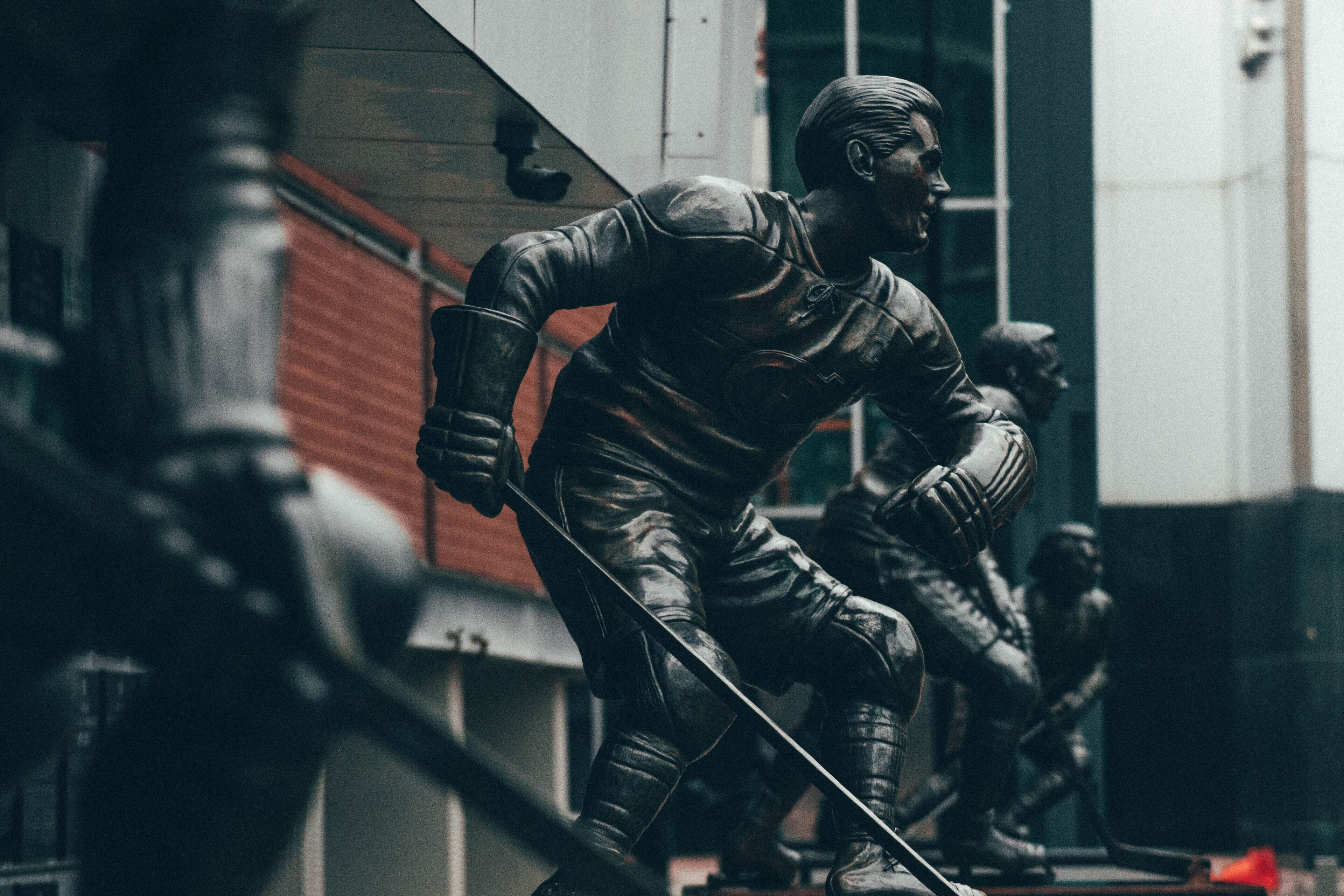 A statue of a person in a leather outfit holding a sword, set against a building with a red brick wall and a glass window.