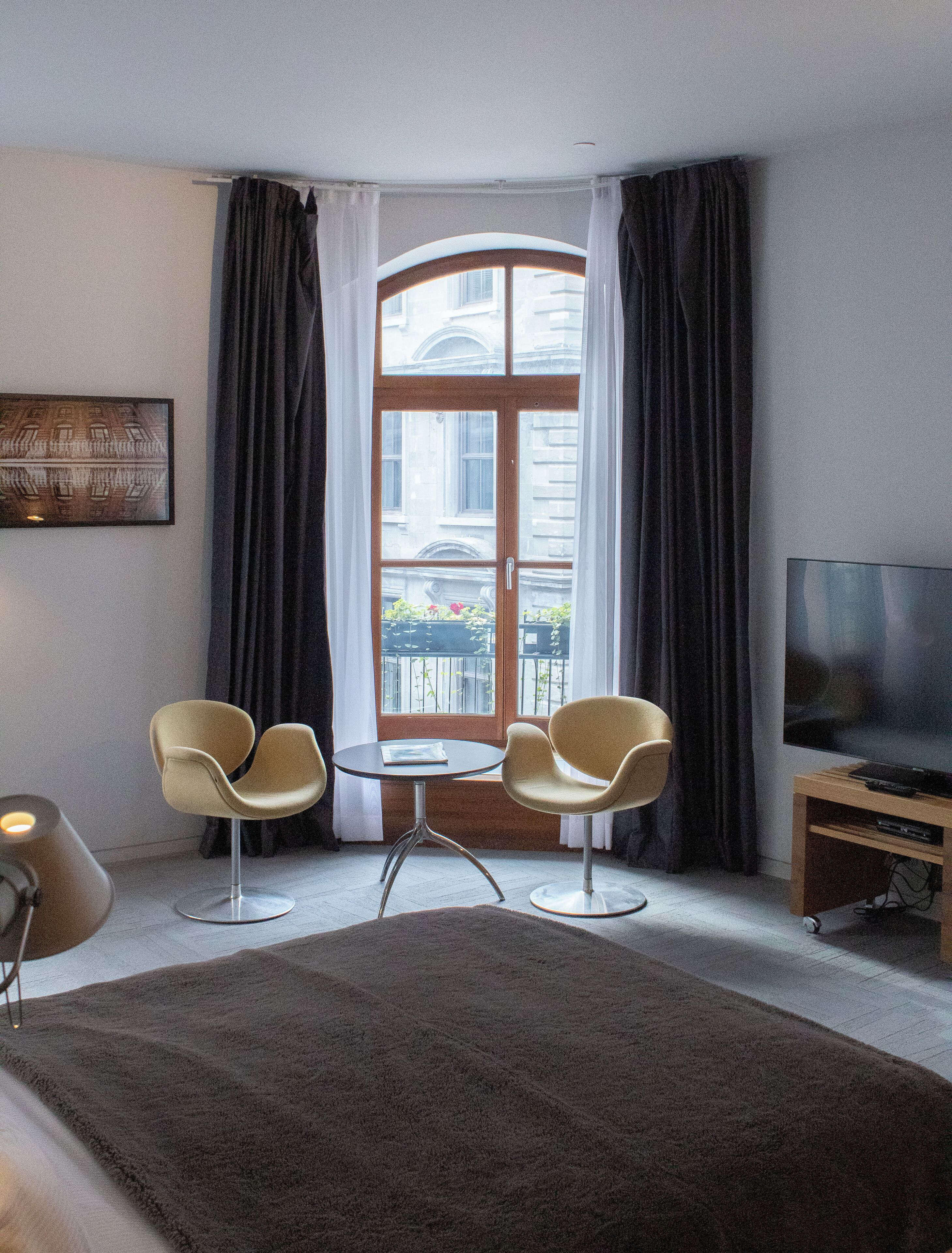 Een hotelkamer met een groot bed, twee fauteuils, een kleine ronde tafel, een televisie op een houten dressoir en een groot raam met gordijnen.
