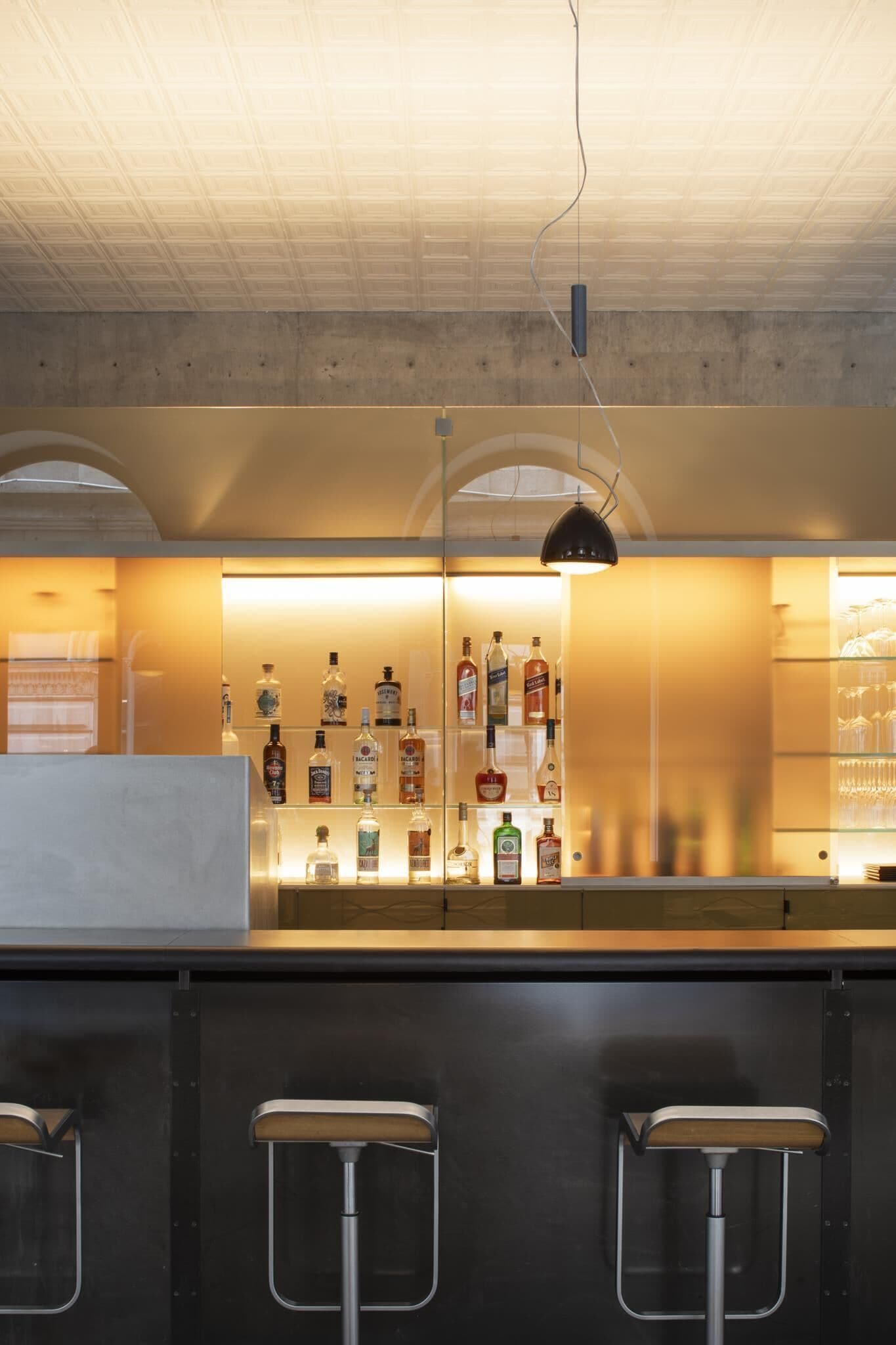 Un espace bar avec un comptoir où sont exposées différentes bouteilles et différents verres, trois tabourets de bar en métal et un luminaire suspendu.