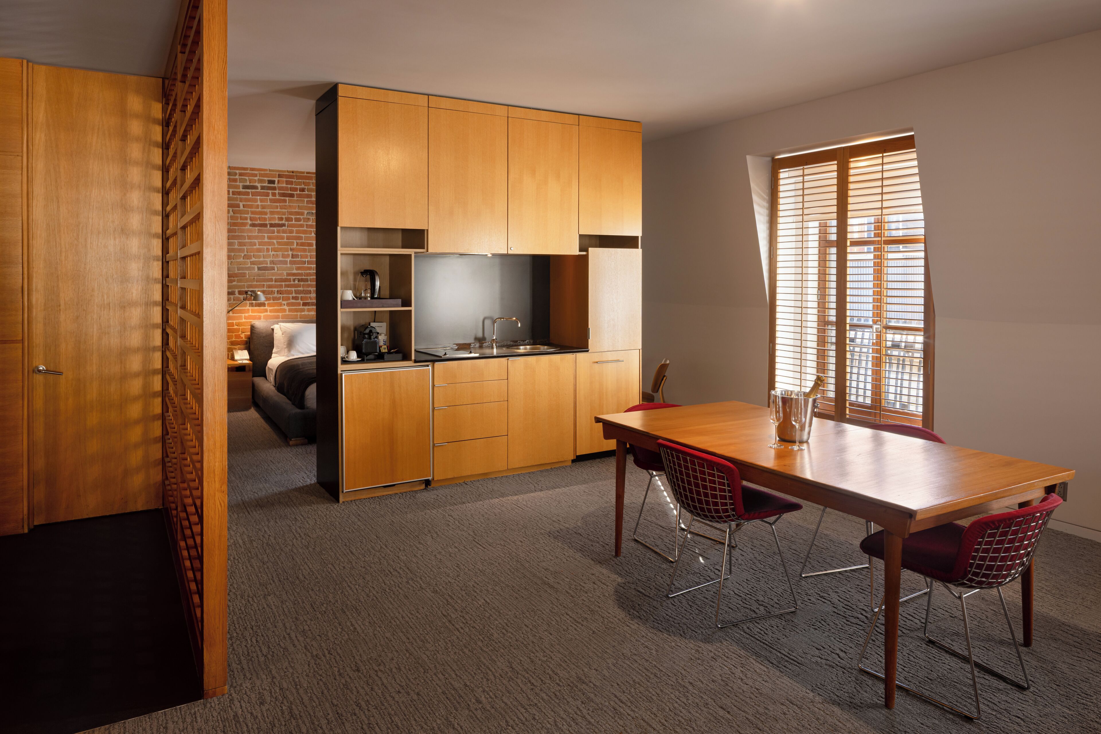 Cocina compacta con armarios de madera, comedor con mesa de madera y sillas rojas, y pared de ladrillo al fondo.