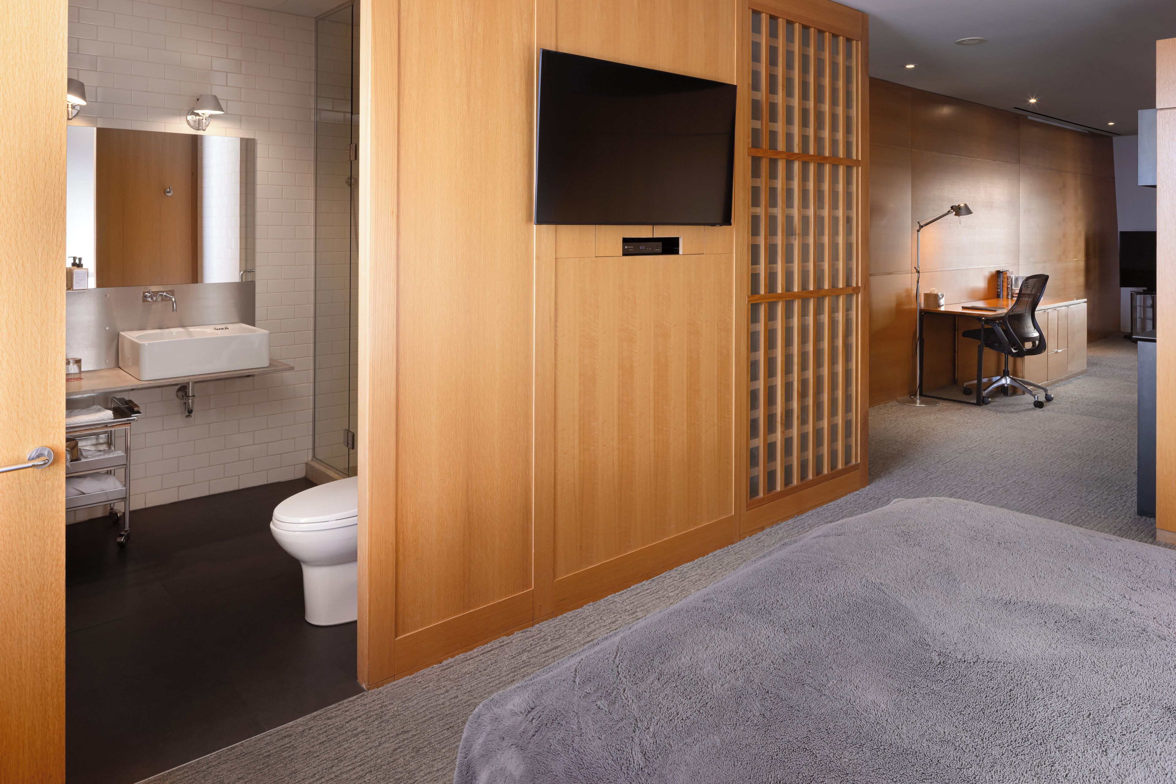 Habitación de hotel moderna con separador de madera, cama grande, escritorio con silla, televisión y baño con lavamanos e inodoro.