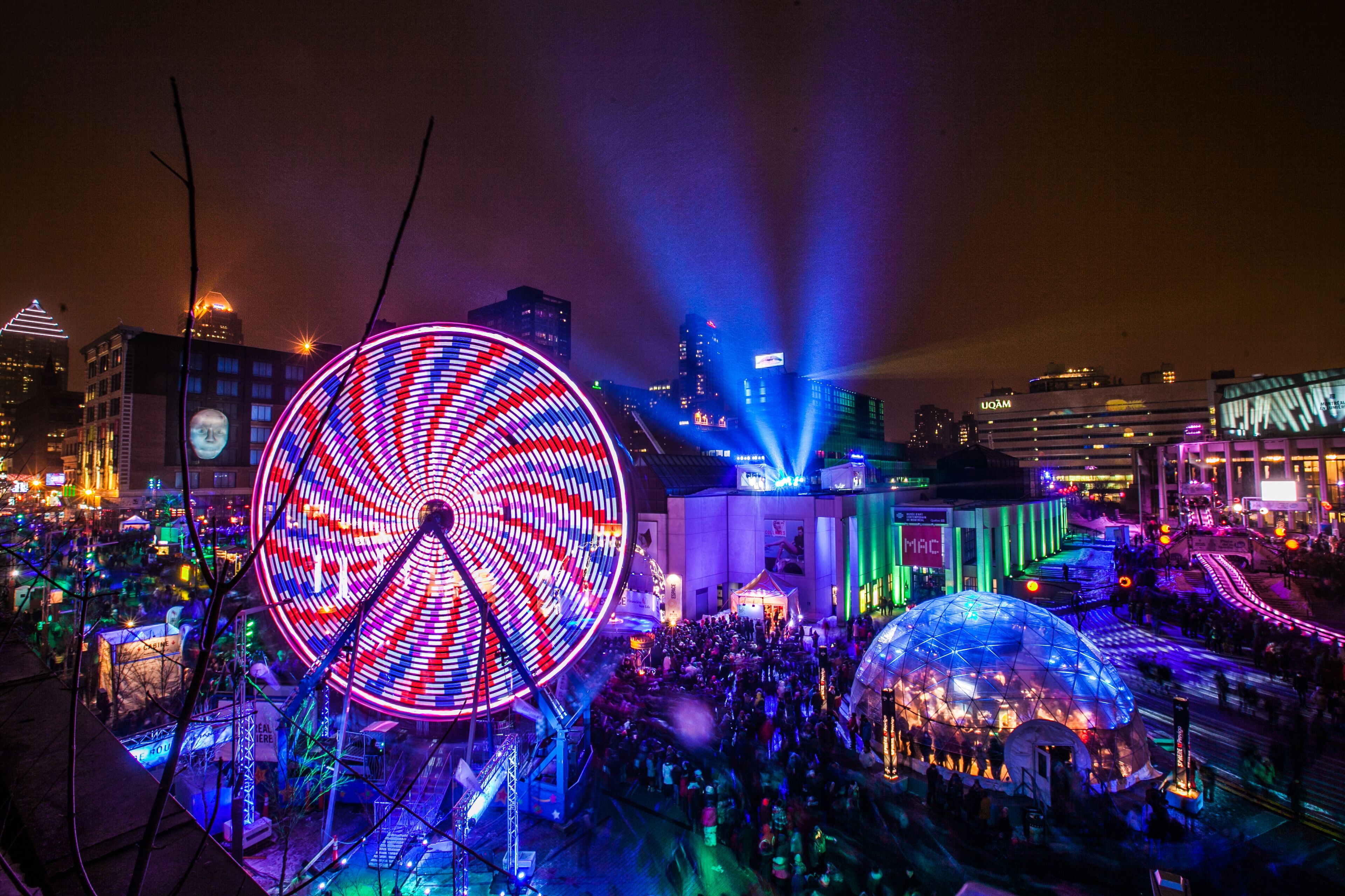 A large Ferris wheel and a dome-shaped structure in a cityscape at night.