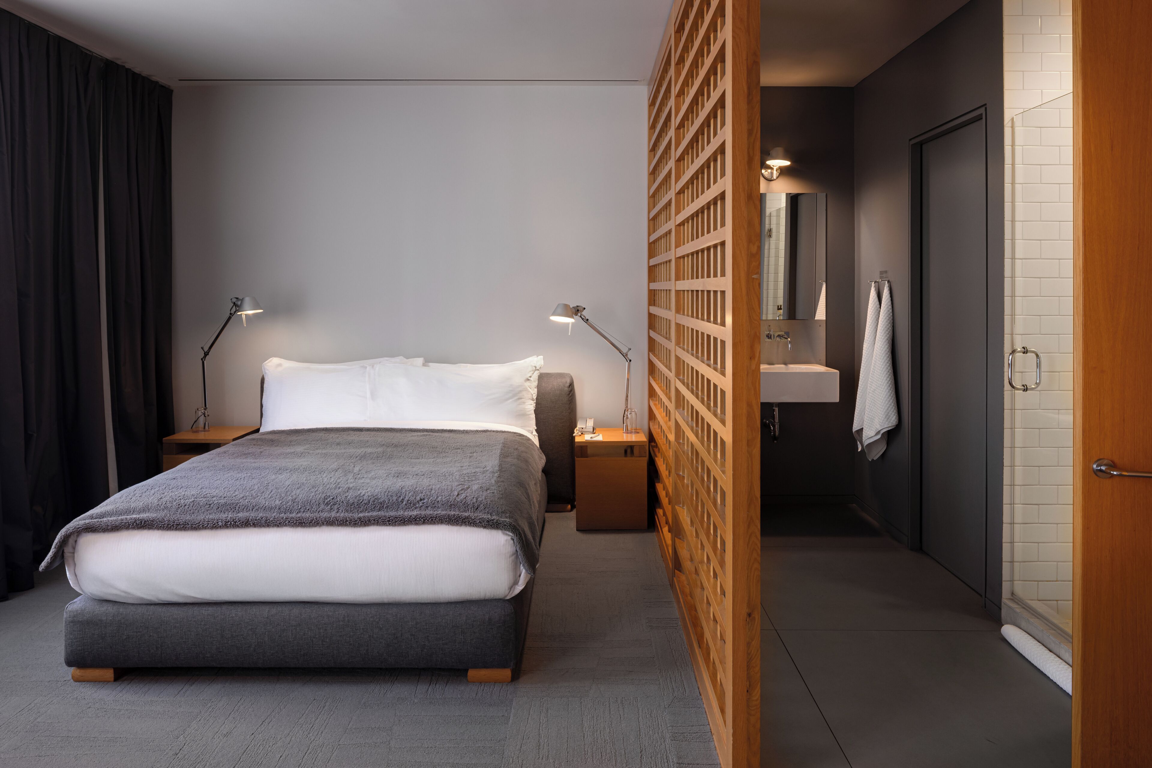 A modern bedroom with a bed, bedside lamps, a wooden partition, a bathroom with a sink and mirror, and a door.