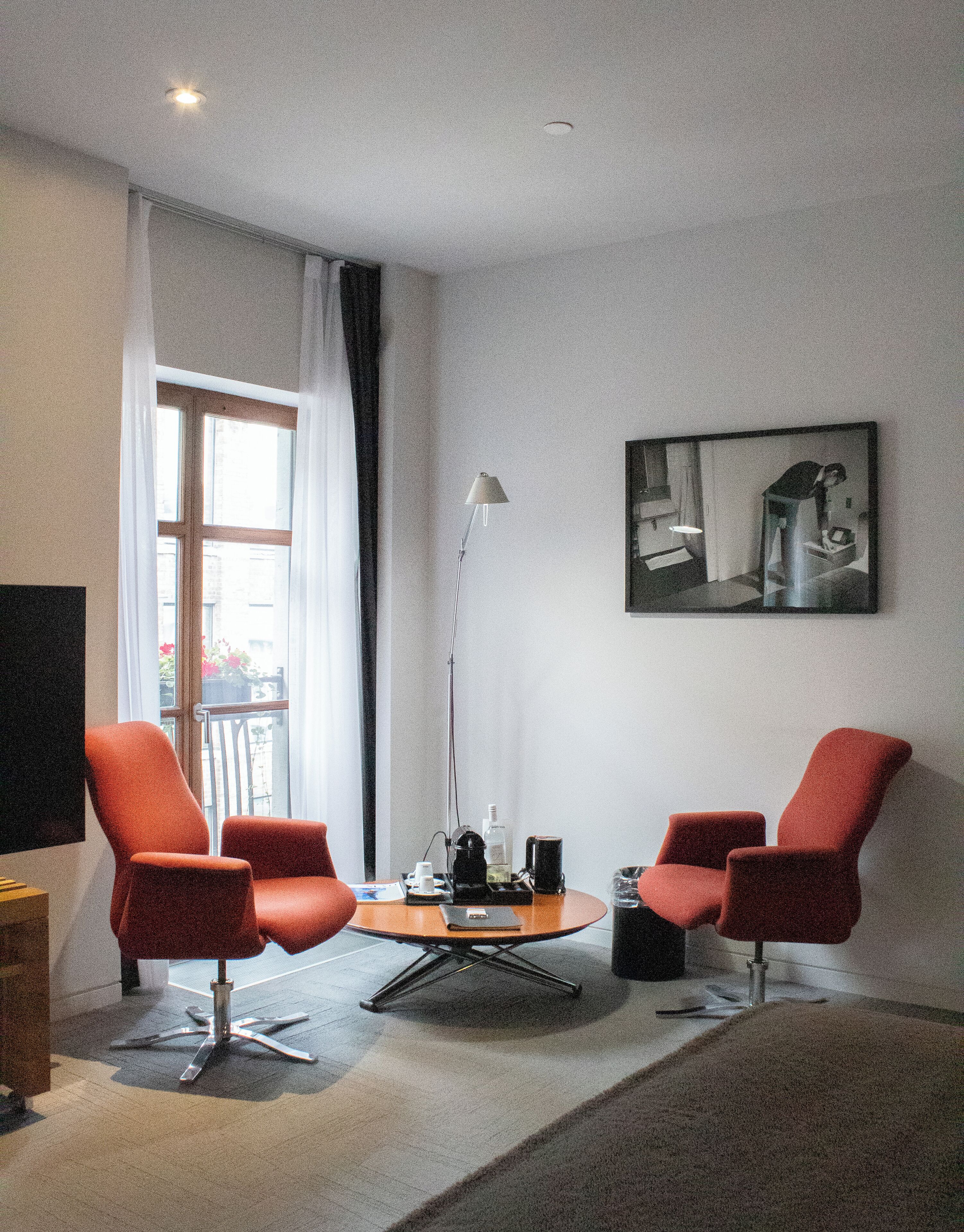 Un salon moderne avec deux fauteuils rouges, une table basse ronde, un lampadaire et un tableau accroché au mur.
