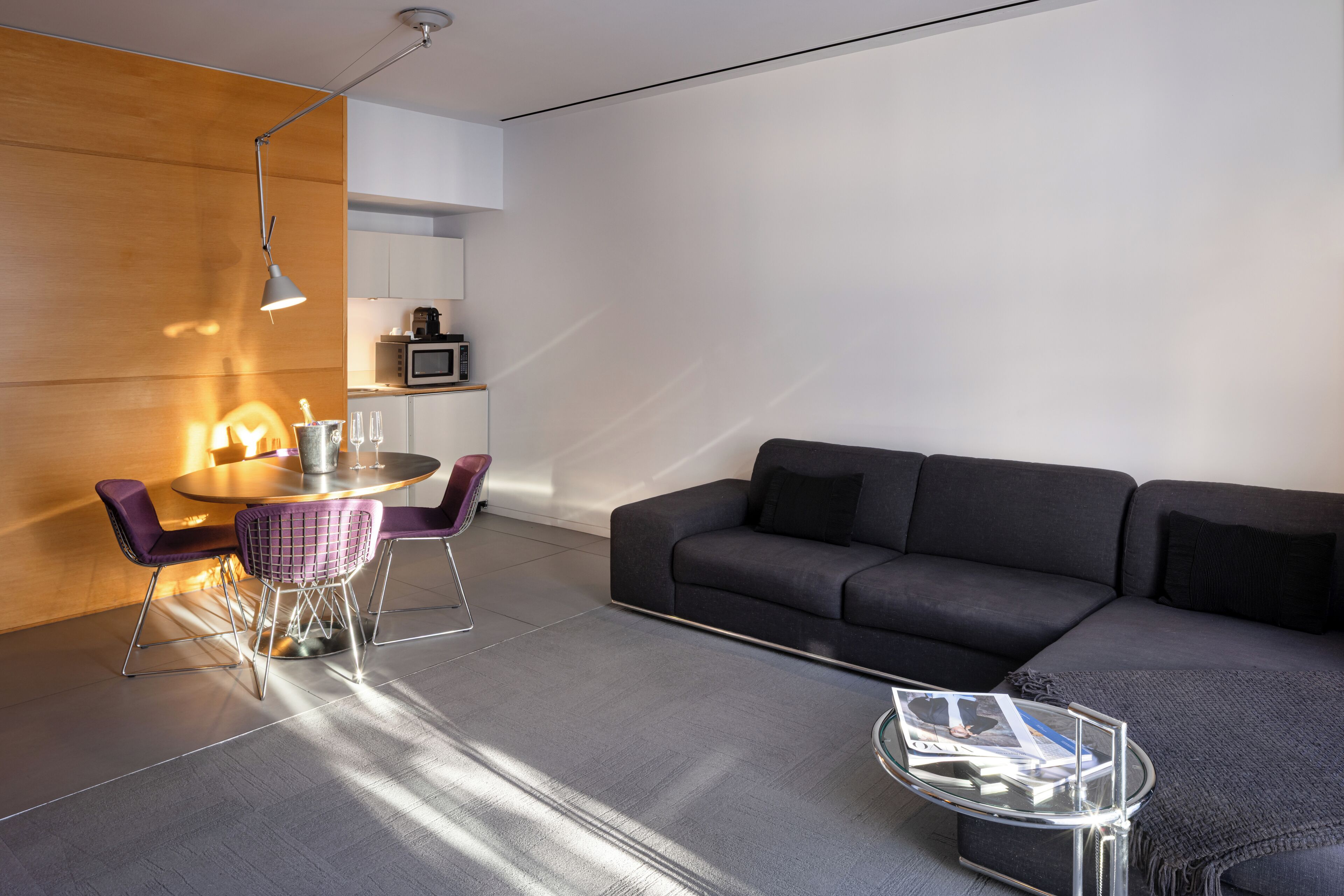 A modern living room with a grey sofa, a round dining table with purple chairs, a glass coffee table, and a pendant light.