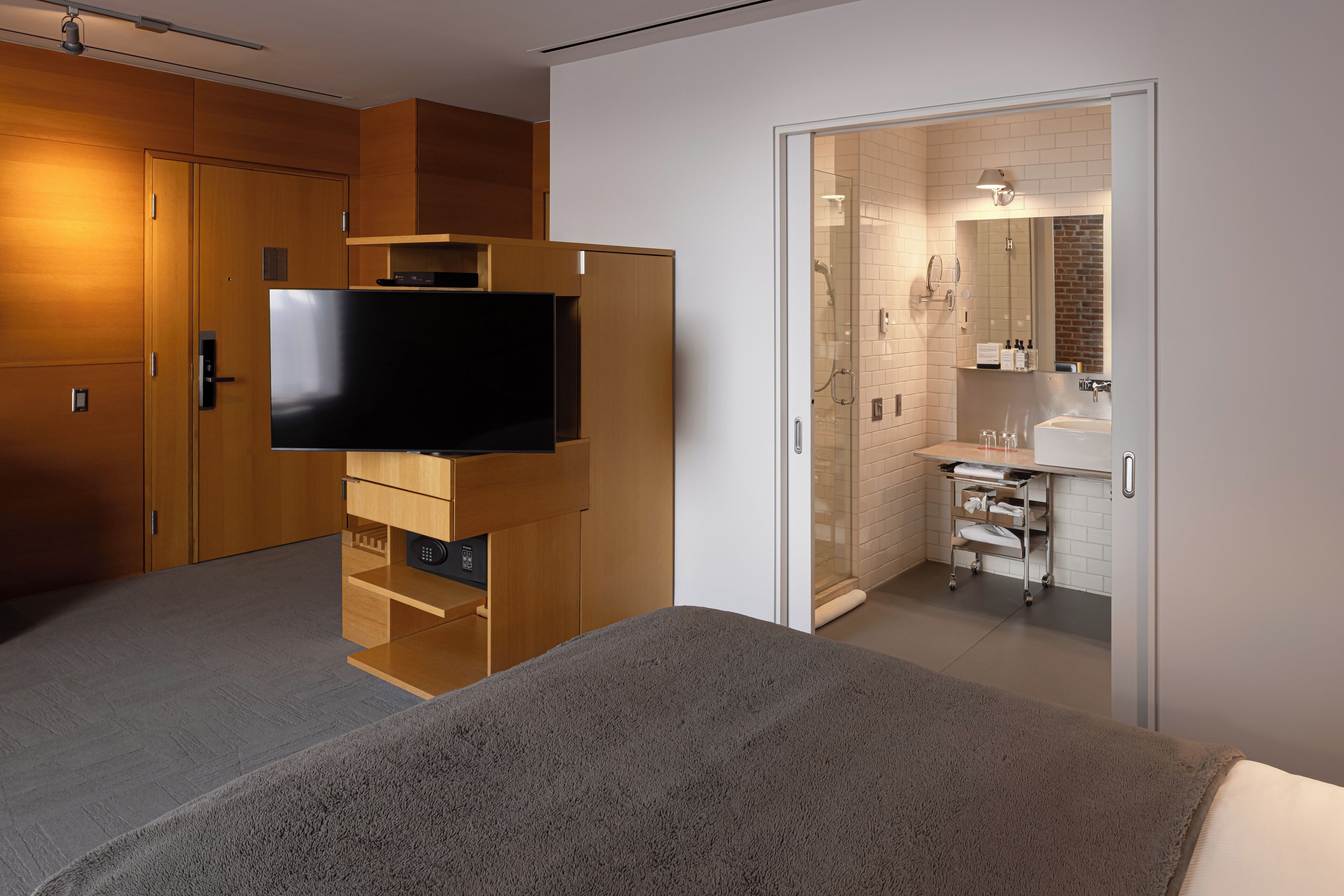 Habitación de hotel con cama, televisor montado en un mueble de madera, baño con lavamanos y espejo, y zona de ducha.