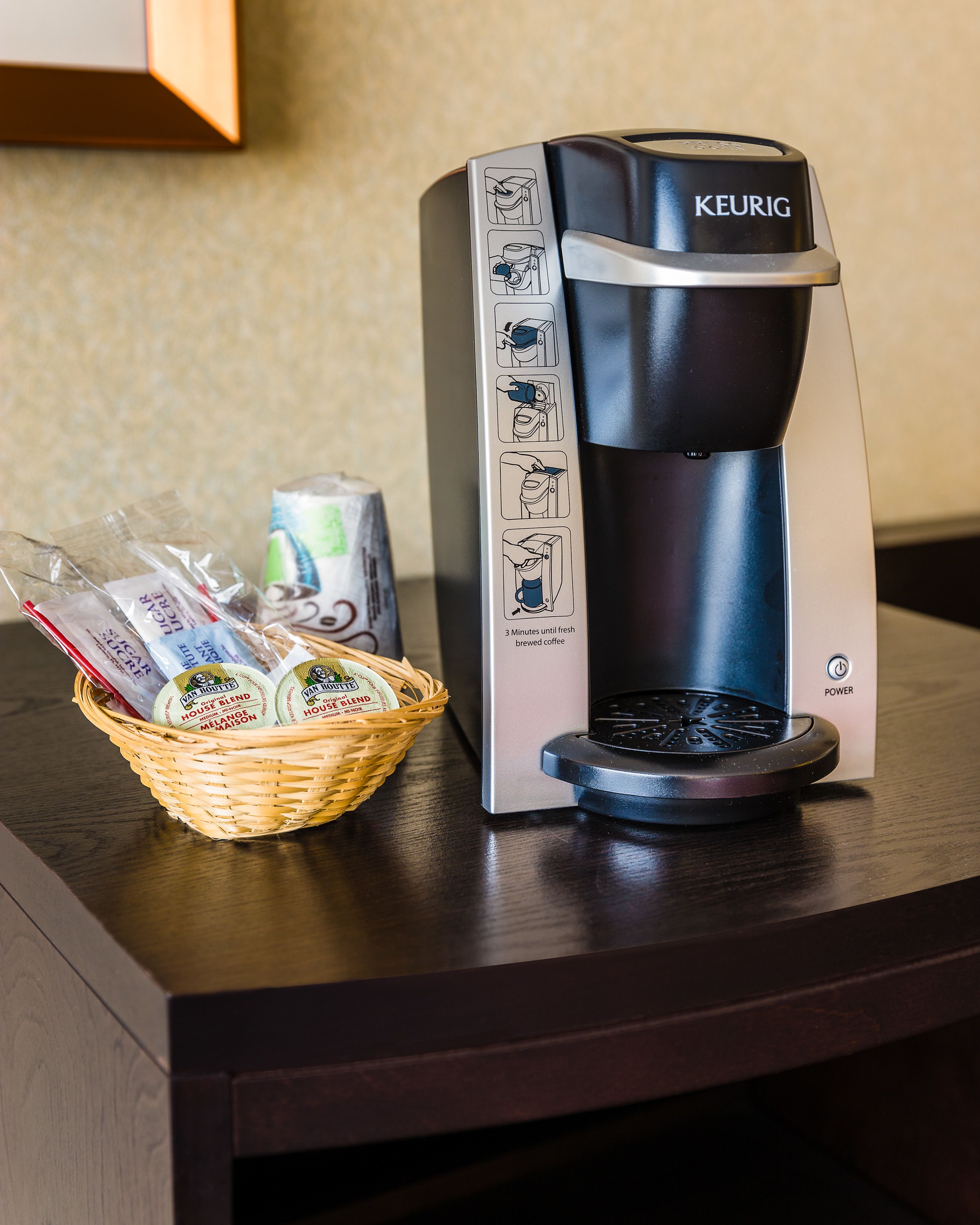 Una cafetera Keurig sobre una taula de fusta amb un cistell de càpsules de cafè i una llauna de refresc.