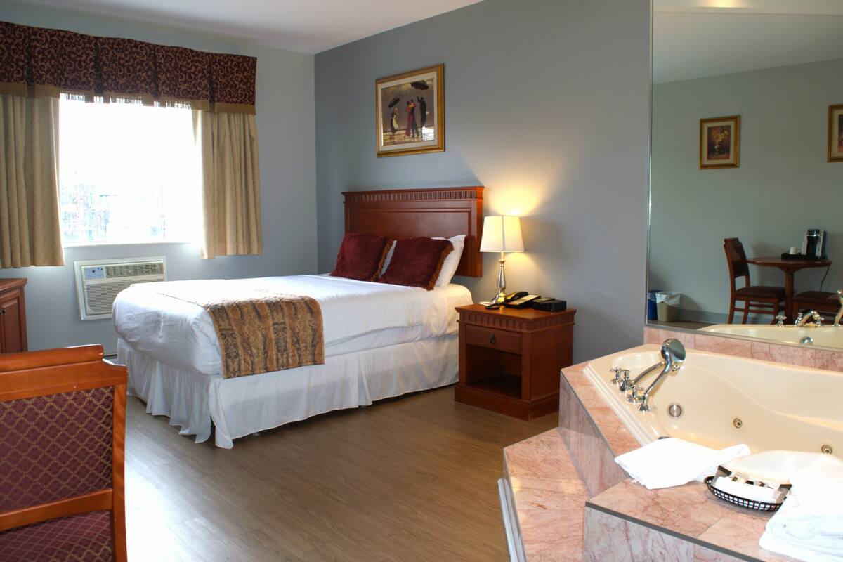 A hotel room with a large bed, a wooden nightstand with a lamp, a bathroom with a bathtub, and a mirror.