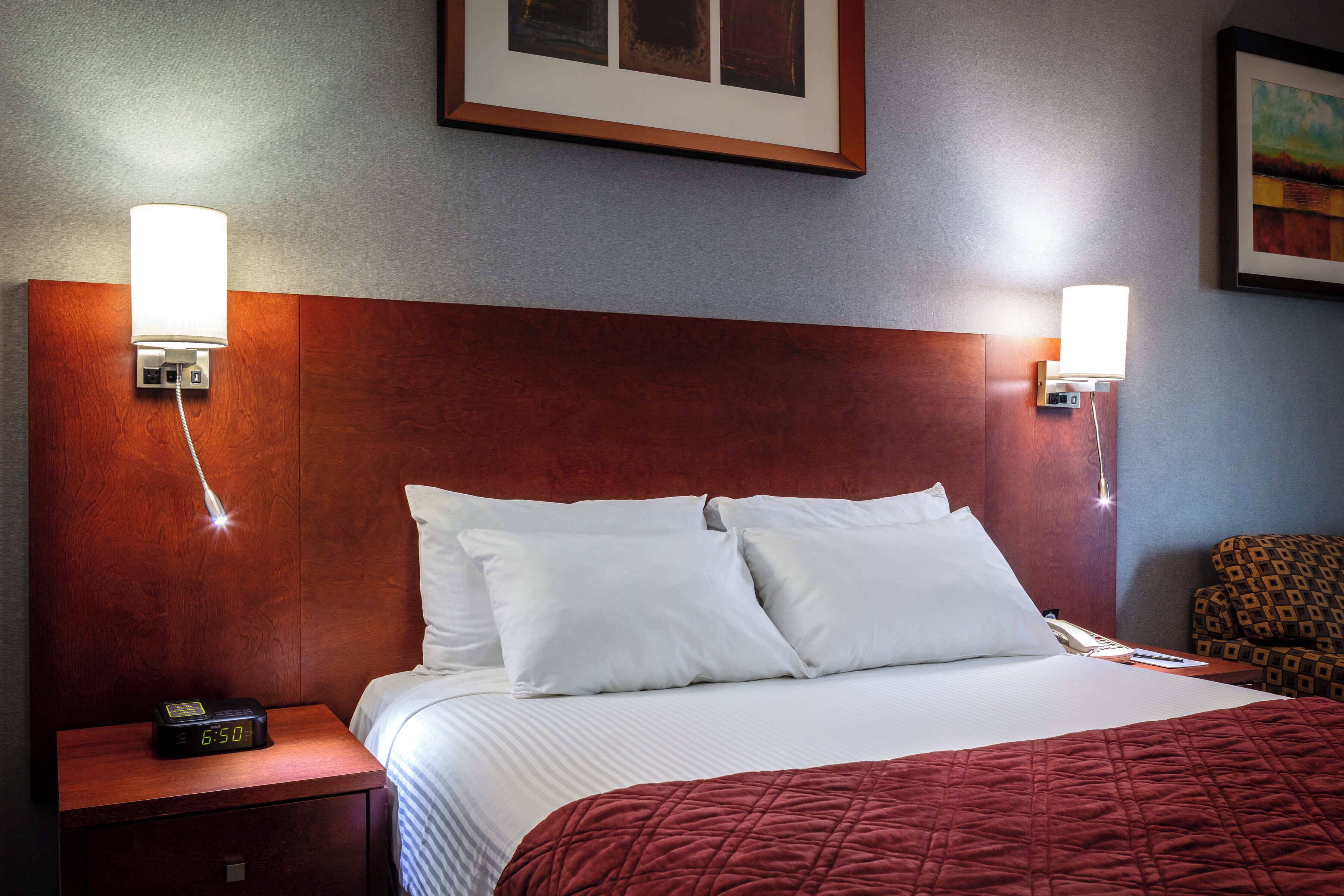 A hotel room with a wooden headboard, a bed with white linens, a red quilted blanket, a nightstand with a clock, and two wall-mounted lamps.