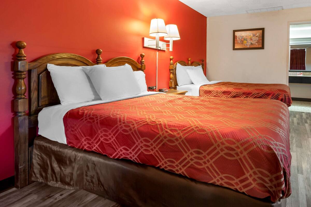 Une chambre d’hôtel avec deux lits, chacun doté d’une tête de lit en bois, de draps blancs et d’une couvre-lit à motifs rouges. Il y a une table de chevet avec une lampe entre les deux lits, et un tableau au mur.