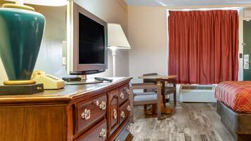 A hotel room with a wooden dresser, a television, a desk with a chair, a red curtain, and an air conditioning unit.