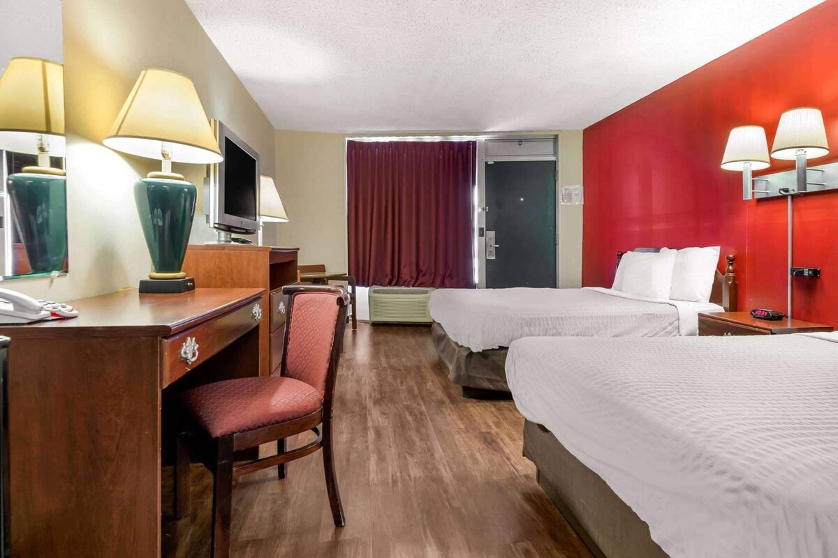 A hotel room with a desk, two beds, a chair, a TV, and a red accent wall.