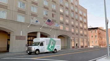 Un camión de reparto blanco estacionado frente a un hotel con varias banderas, incluyendo la bandera estadounidense, y un edificio grande de diseño moderno.