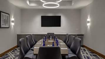 A conference room with a long table, black leather chairs, a flat-screen TV, and a patterned carpet.