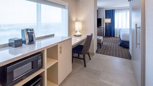 A hotel room with a kitchenette, a desk with a lamp, a chair, a bed with blue curtains, and a TV. - Embassy Suites Boston Logan Airport (Boston)