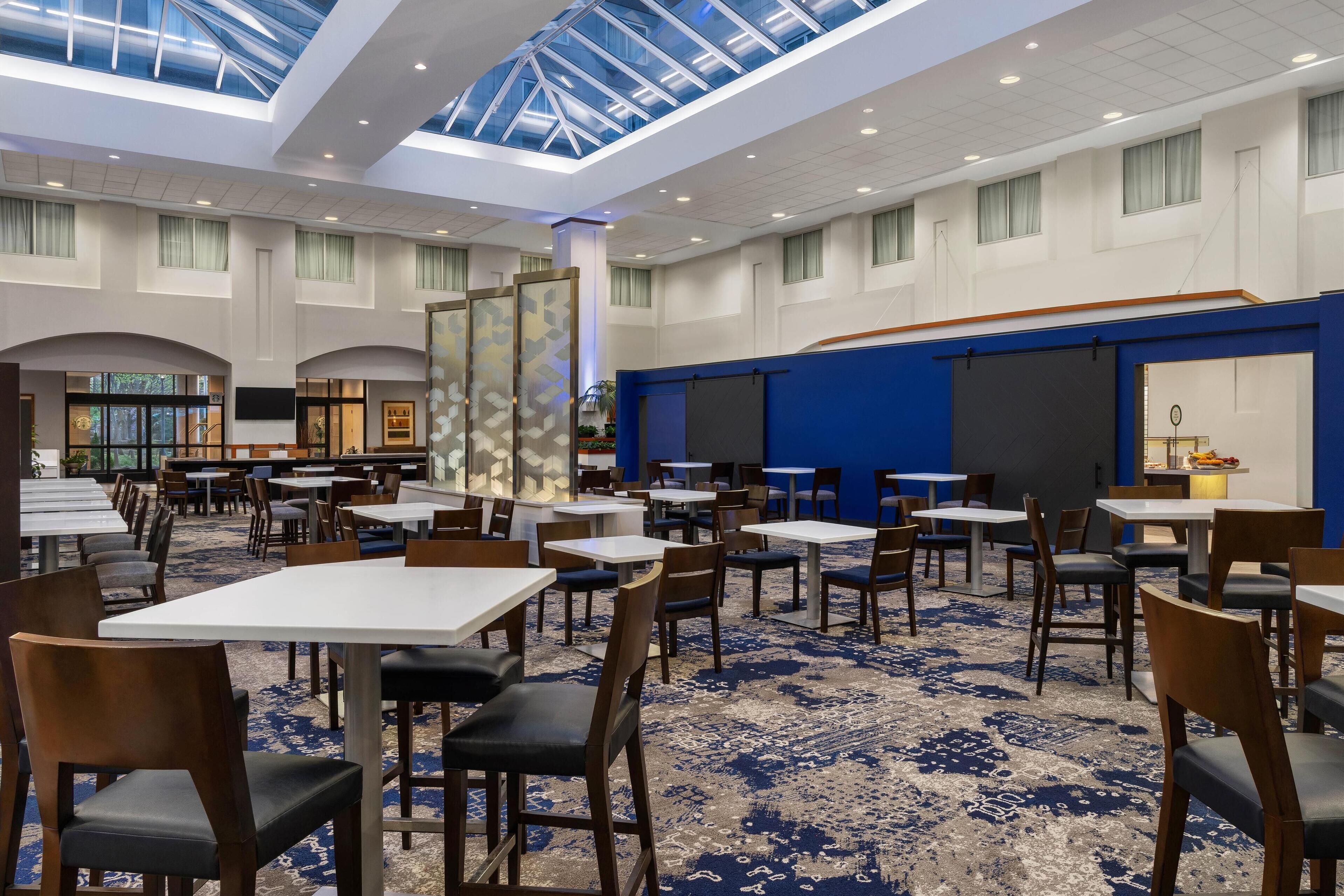 A spacious hotel dining area with multiple tables and chairs, a large glass ceiling, and a blue accent wall.