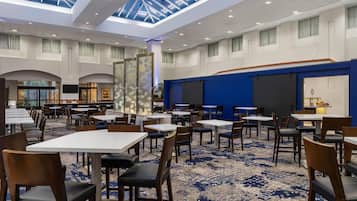 A spacious hotel dining area with multiple tables and chairs, a large glass ceiling, and a blue accent wall.