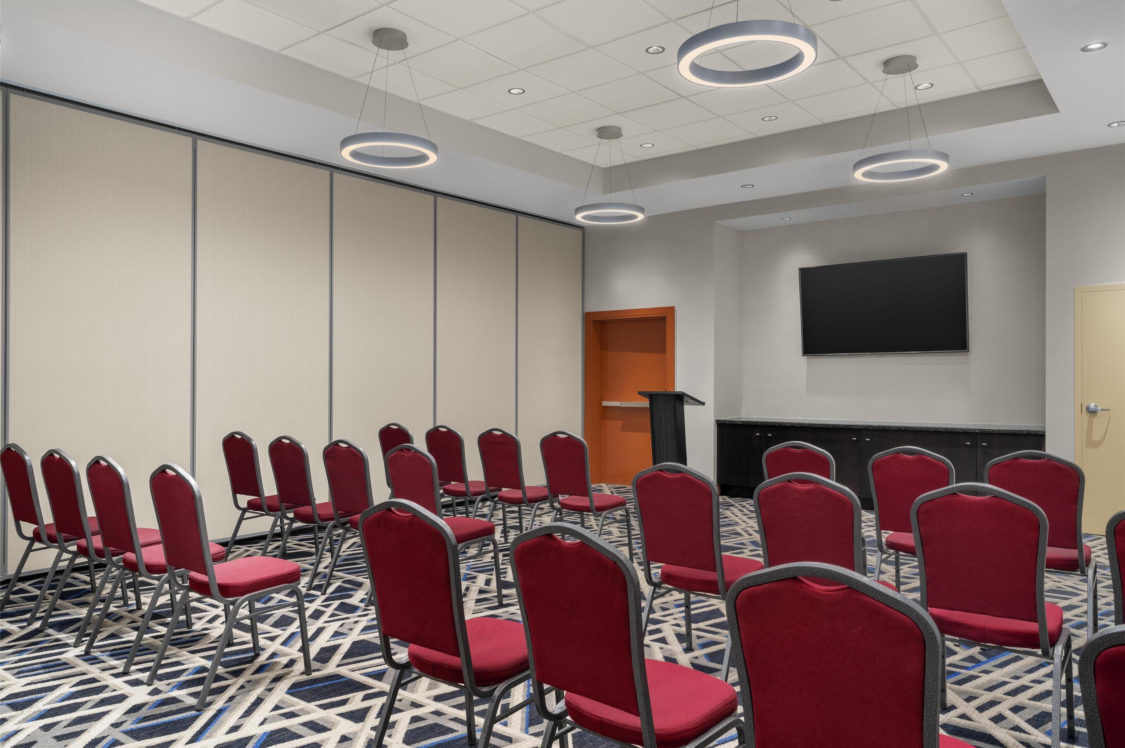 Une salle de conférence avec des rangées de chaises rouges, un pupitre et un grand écran mural.