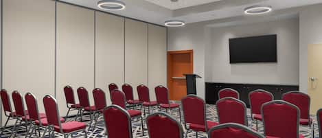 Une salle de conférence avec des rangées de chaises rouges, un pupitre et un grand écran mural.