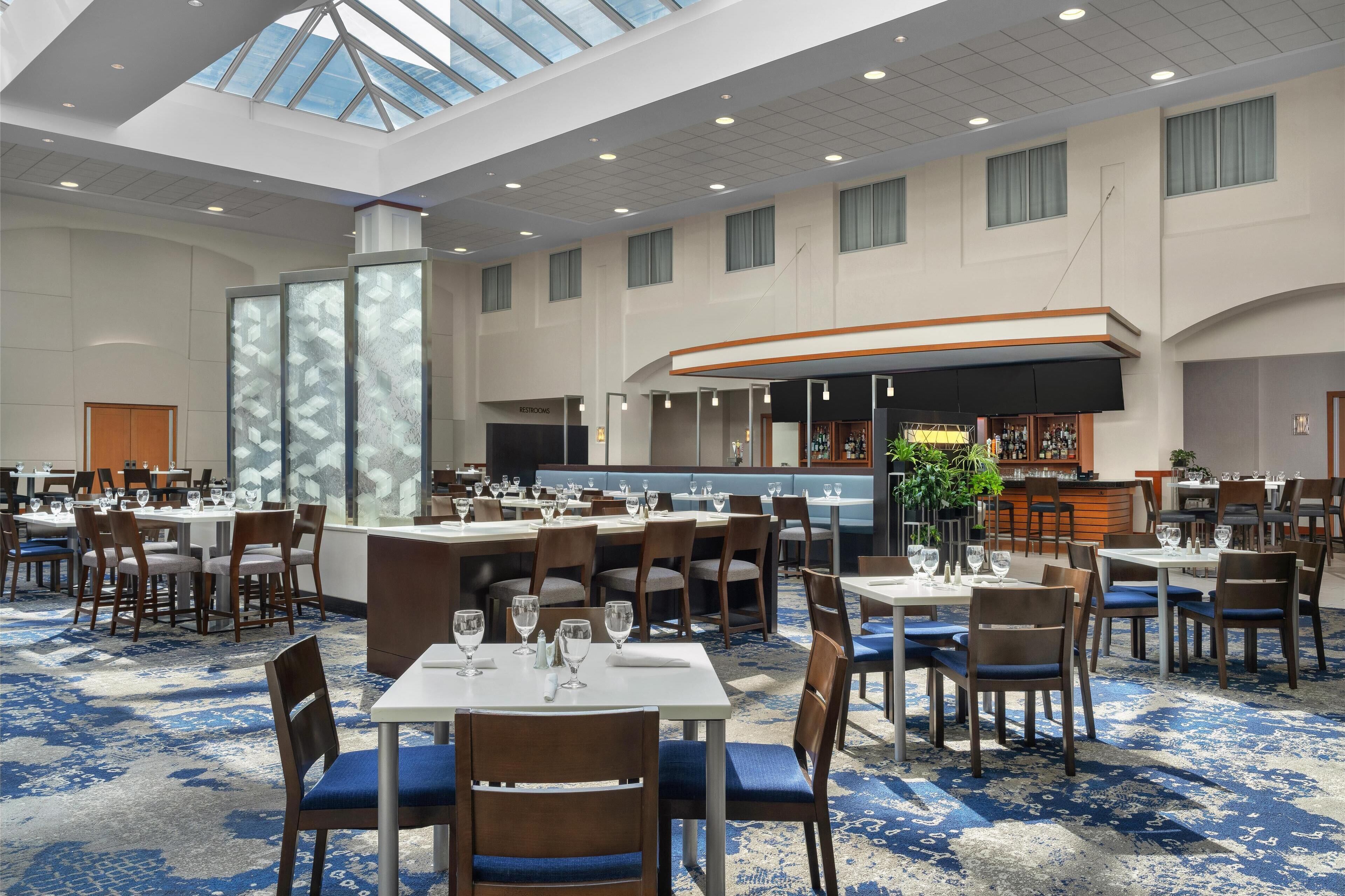 Un vaste espace de restauration dans l’hôtel, avec de nombreuses tables et chaises, un bar et une grande fenêtre laissant entrer la lumière naturelle.
