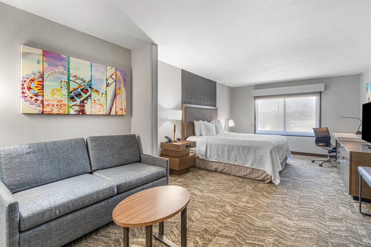 Habitación de hotel con un sofá gris, una cama con edredón blanco, un escritorio con silla y un ventanal grande.