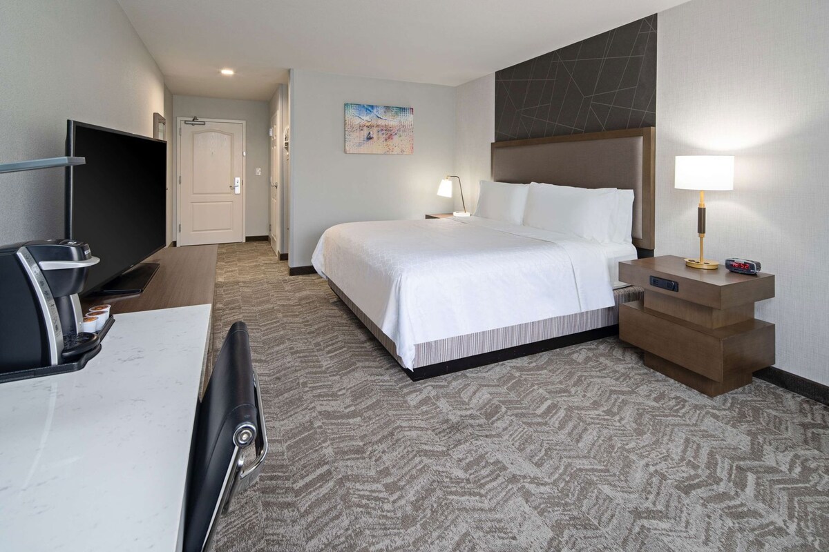 A hotel room with a bed, bedside table, lamp, TV, and coffee maker.