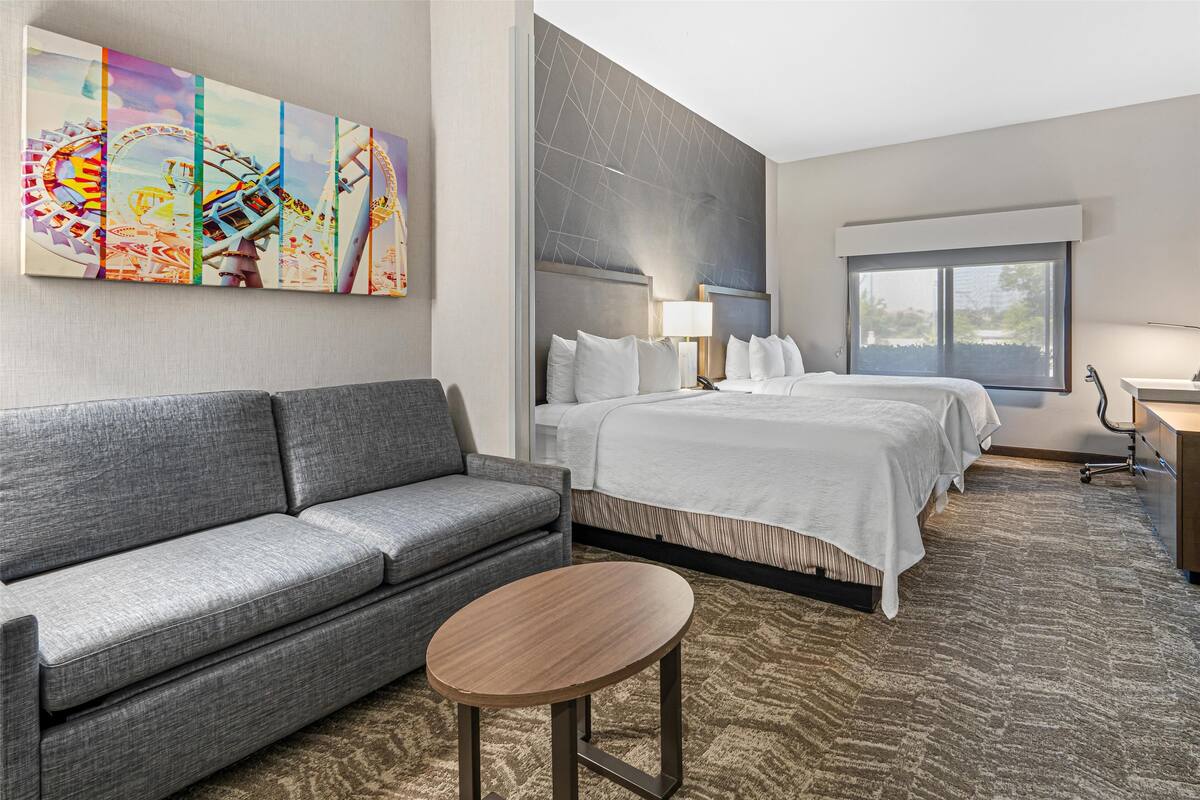 Habitación de hotel con sofá gris, cama individual con ropa de cama blanca, mesita redonda pequeña, ventana grande y un cuadro colgado en la pared que representa una escena de carnaval.