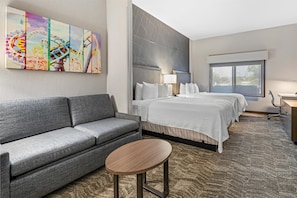 A hotel room with a gray sofa, a single bed with white bedding, a small round table, a large window, and a wall-mounted artwork depicting a carnival scene. - Best Western Valencia/Six Flags Inn & Suites (Valencia)