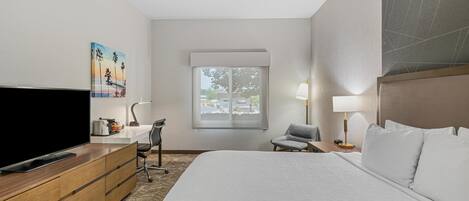 A hotel room with a large bed, a flat-screen TV, a desk with a lamp, a chair, and a window with a view of the outside.