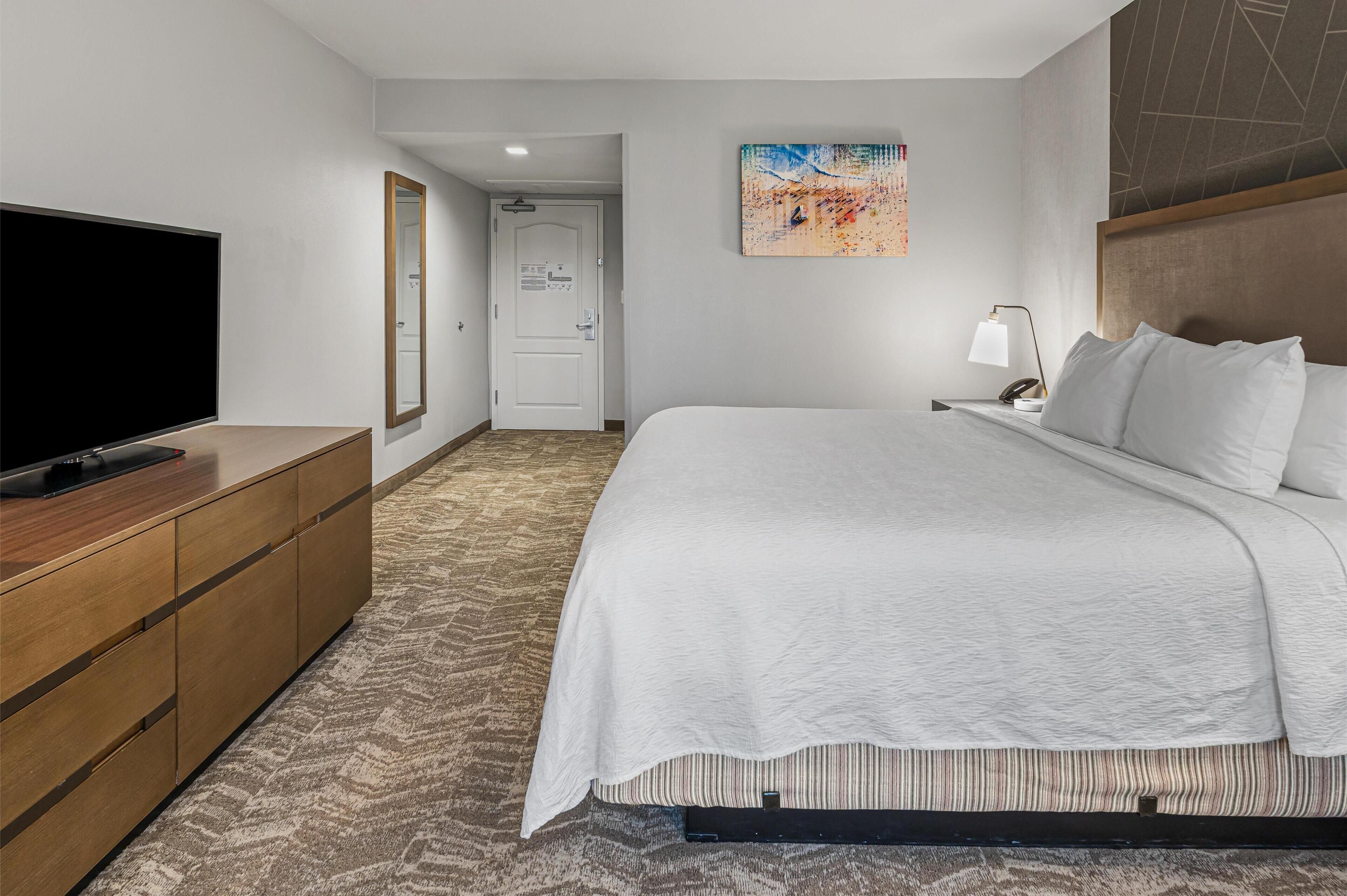Ein Hotelzimmer mit einem großen Bett, einem Flachbildfernseher auf einer Holzkommode, einem Bild an der Wand und einer Tür zu einem weiteren Raum.
