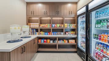A kitchenette with a refrigerator stocked with beverages, a coffee machine, and shelves with snacks and condiments.